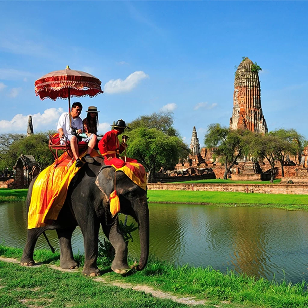 Tourists riding an elephant dressed in traditional Thai attire near a serene river, with ancient Ayutthaya temples in the background under a bright blue sky.