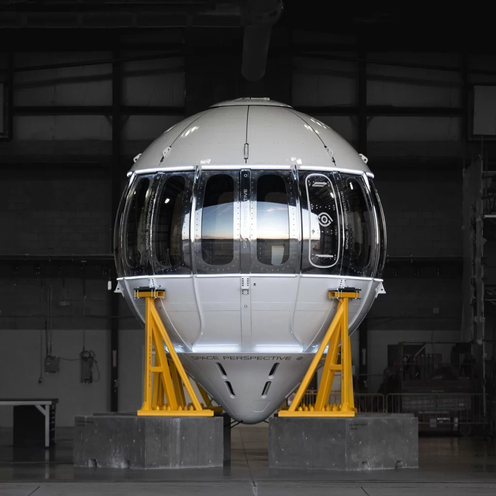 The Spaceship Neptune capsule, a sleek, spherical spacecraft with large panoramic windows, displayed inside a hangar.