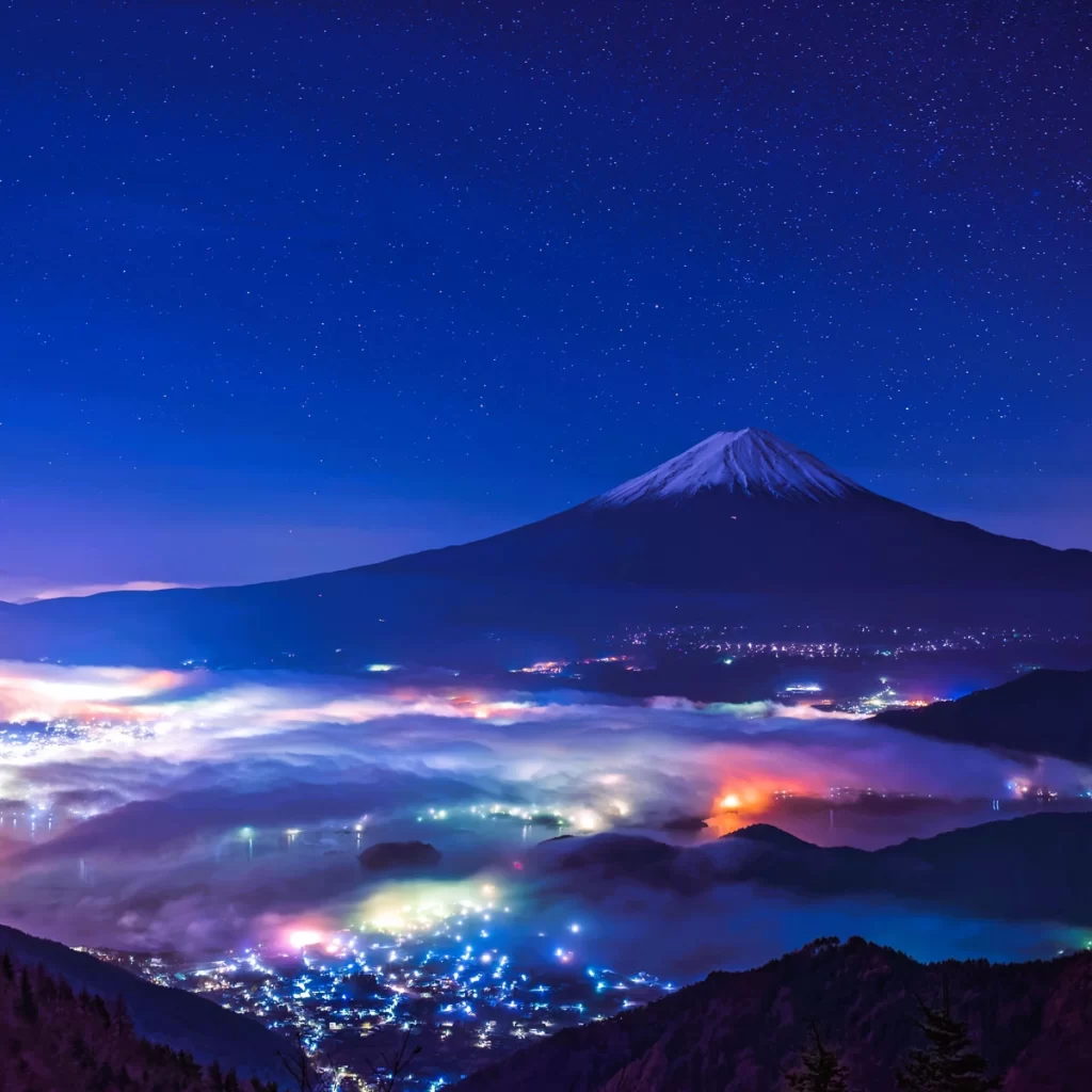 A serene view of Mount Fuji at night, with a snow-capped peak rising above vibrant, mist-covered city lights in the valley below