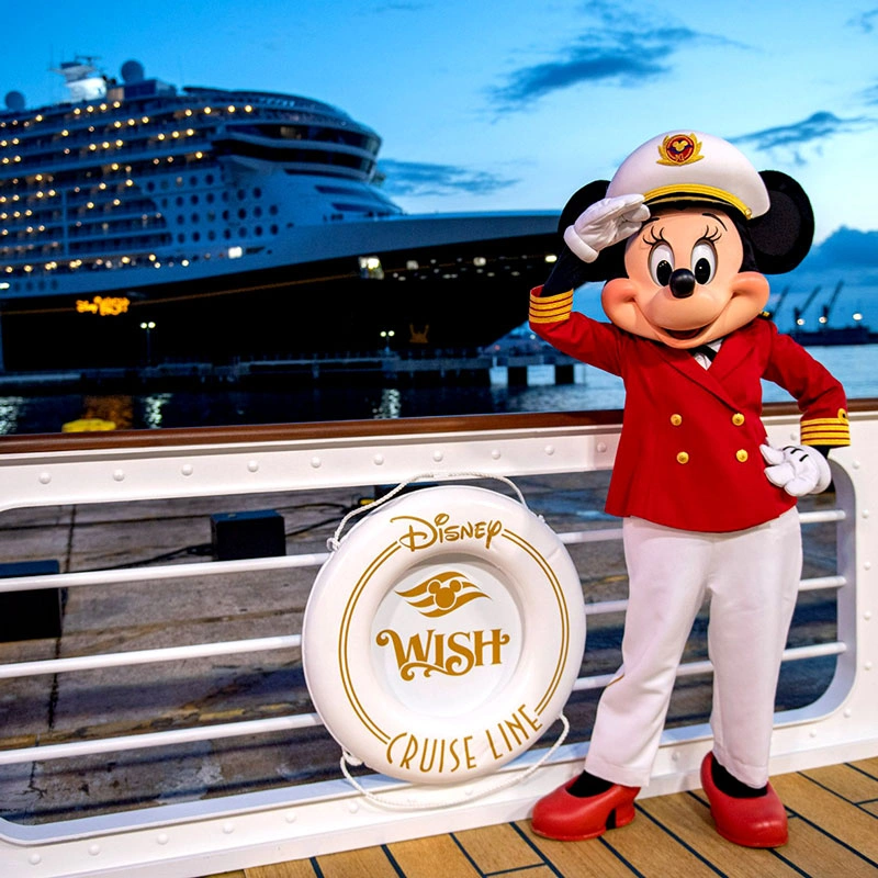 Minnie Mouse dressed as a ship captain saluting on the deck of a Disney Cruise Line ship during sunset, with a lifebuoy labeled "Disney Wish" in the foreground.