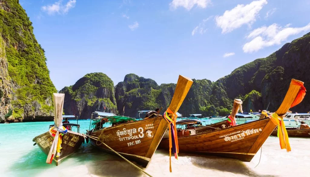 A panoramic view of Railay Beach in Krabi, Thailand, with turquoise waters, white sand, and towering limestone cliffs under a bright blue sky