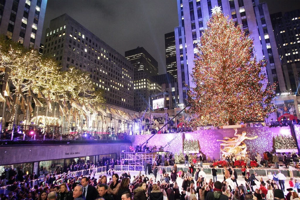 A large, beautifully lit Christmas tree at Rockefeller Center, with people skating at the ice rink below, surrounded by festive lights and decorations