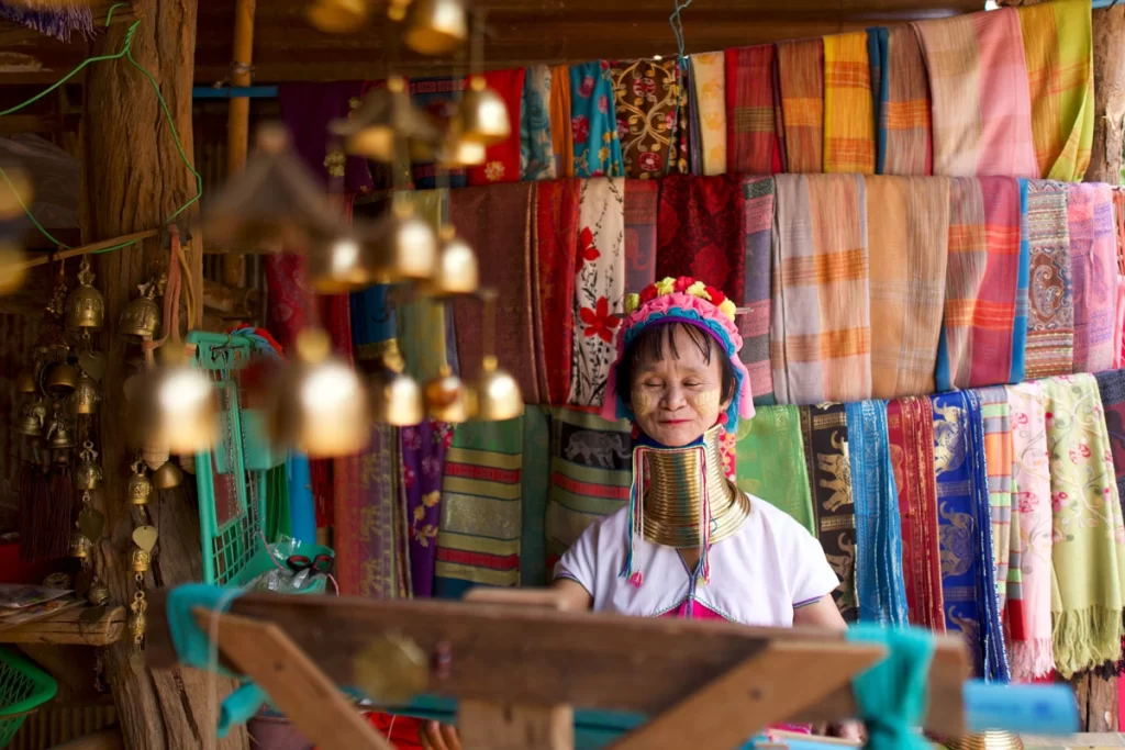 A Karen woman in traditional attire weaving colorful fabric on a handloom in a hill tribe village in Northern Thailand