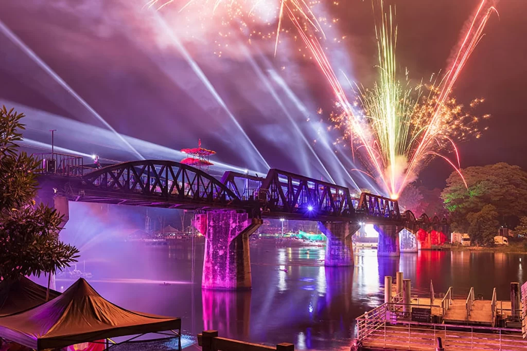 A vibrant night view of the River Kwai Bridge illuminated with colorful lights and fireworks during a festival in Kanchanaburi, Thailand.