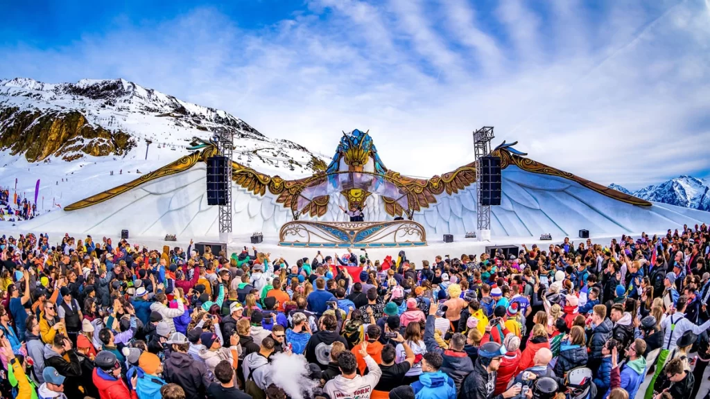 A vibrant Tomorrowland Winter stage surrounded by snowy mountains, with a crowd enjoying the music under colorful lights.