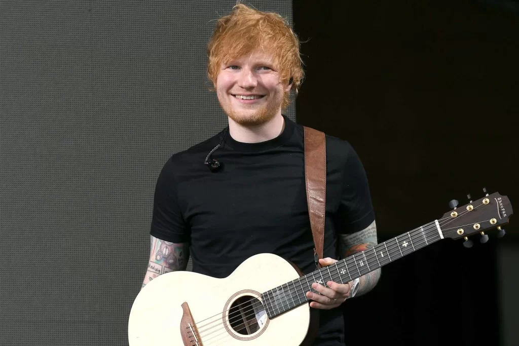 Ed Sheeran holding a guitar, smiling on stage during a live performance.