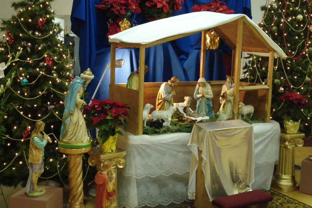 Nativity scene with figures of Mary, Joseph, baby Jesus, and shepherds in a stable setting, surrounded by Christmas trees and festive decorations