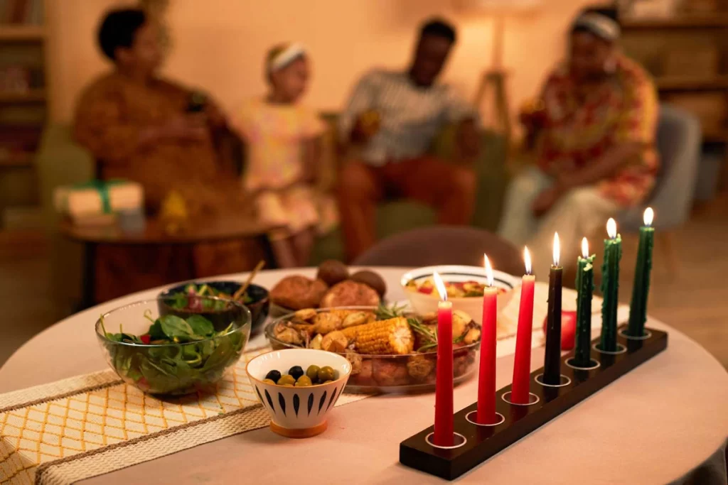 A Kwanzaa celebration table with the Kinara candle holder, holding red, black, and green candles, lit to honor the seven principles, along with traditional food and family members in the background