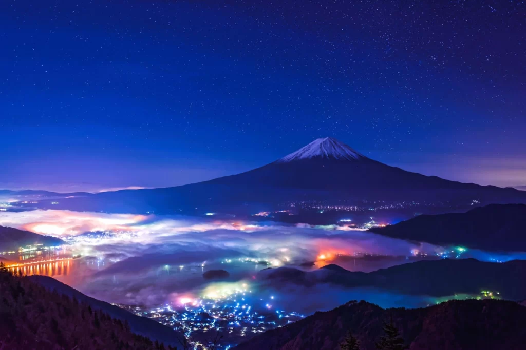 Mount Fuji under a starry night sky, with a snow-capped peak and colorful lights glowing in the misty valleys below