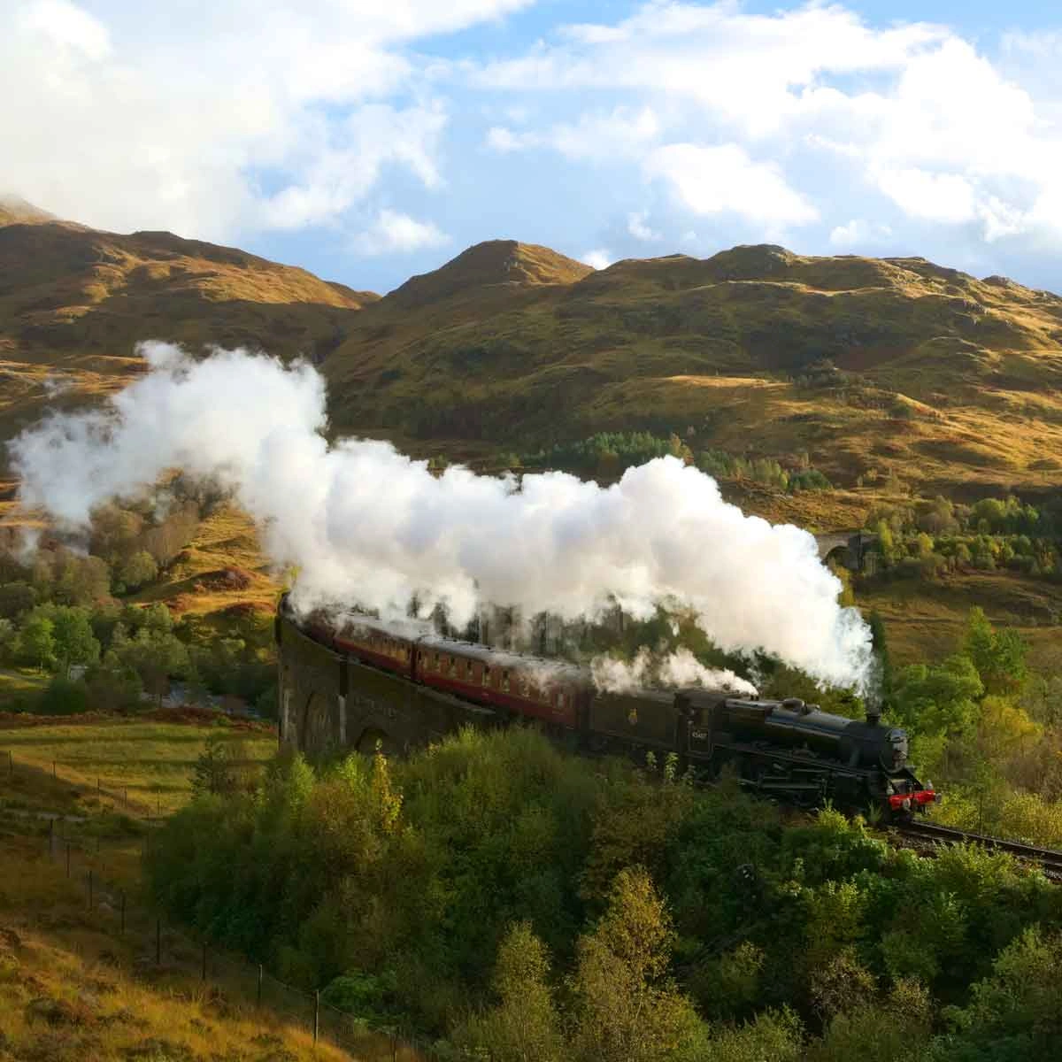 A steam train crossing the Glenfinnan Viaduct in Scotland, surrounded by lush greenery and mountains, known as the real-life Hogwarts Express.