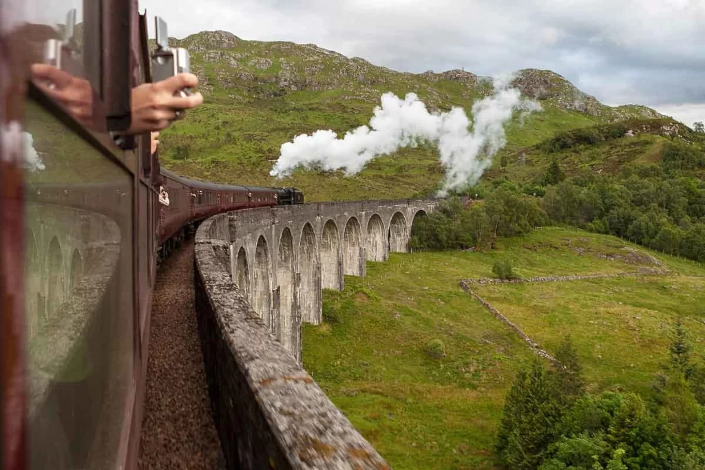 A vintage steam train crossing the Glenfinnan Viaduct, releasing white steam, with lush green hills and a cloudy sky in the background.