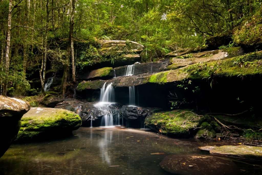 Tham Yai Waterfall cascading over multi-layered rocks surrounded by lush greenery.