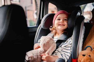 A happy toddler sitting in a forward-facing car seat, securely strapped with a harness, holding a plush toy inside a moving vehicle