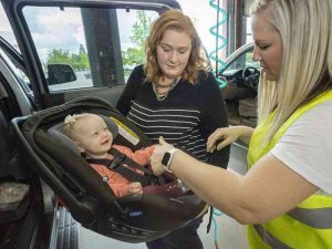 A baby smiling while securely fastened in an infant car seat, with two women assisting in checking the installation inside a vehicle